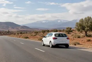 Voiture de location blanche roulant sur une route marocaine avec les montagnes de l’Atlas en arrière-plan.